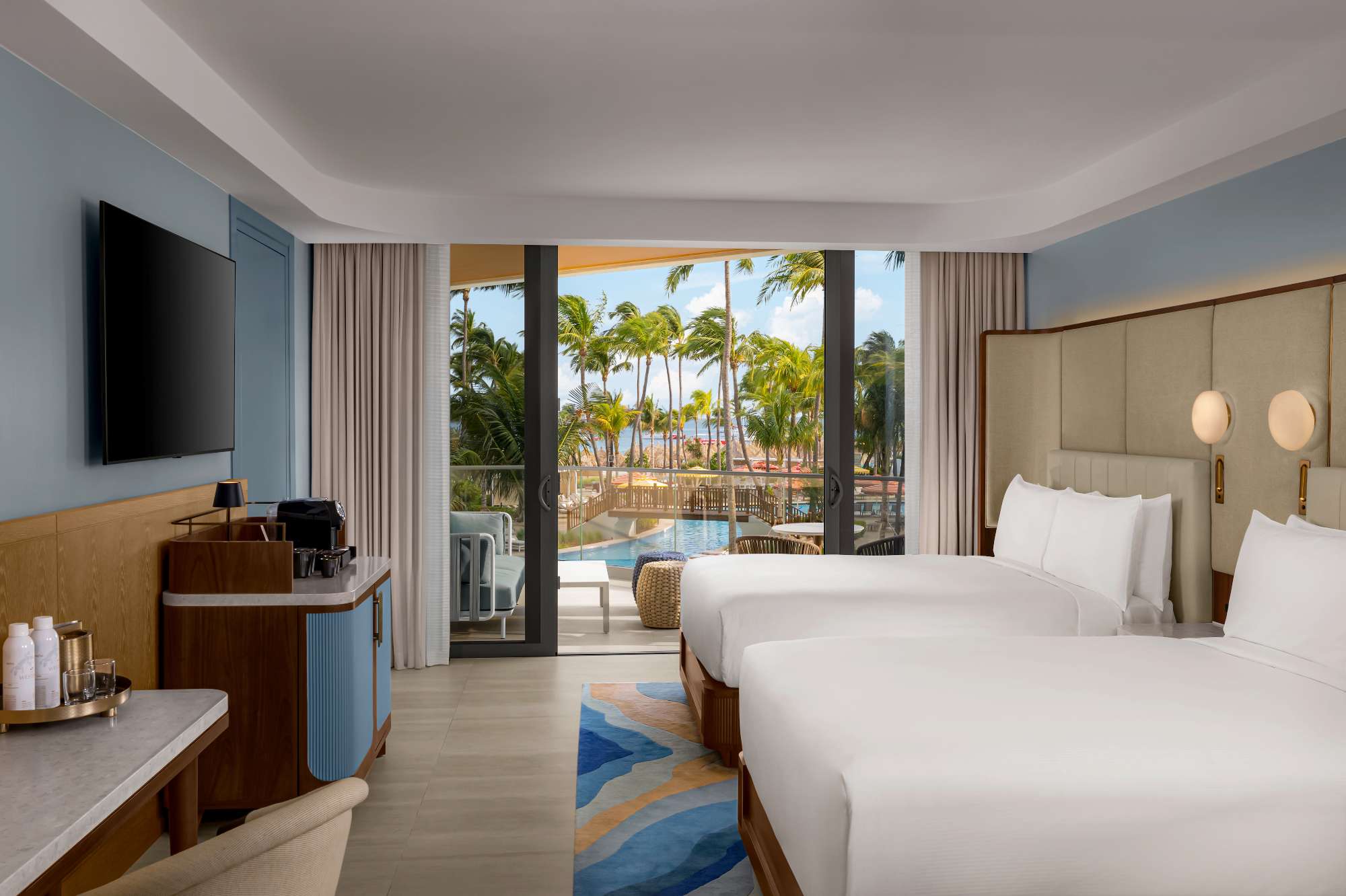 A modern hotel room with two double beds, a TV, a coffee station, and a balcony overlooking a pool and palm trees, with the ocean visible in the background.