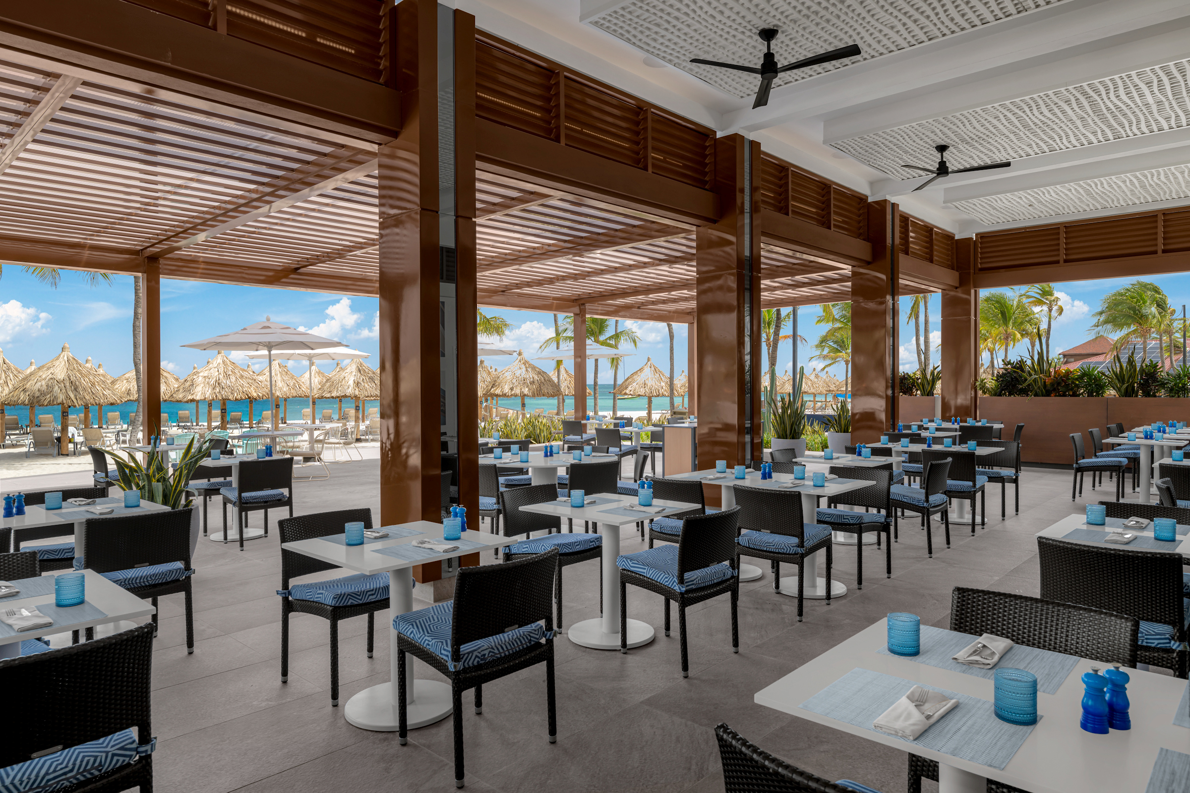 Open-air restaurant with black chairs and white tables, set for dining. Thatched umbrellas, palm trees, and a view of the ocean are visible outside under a sunny sky.