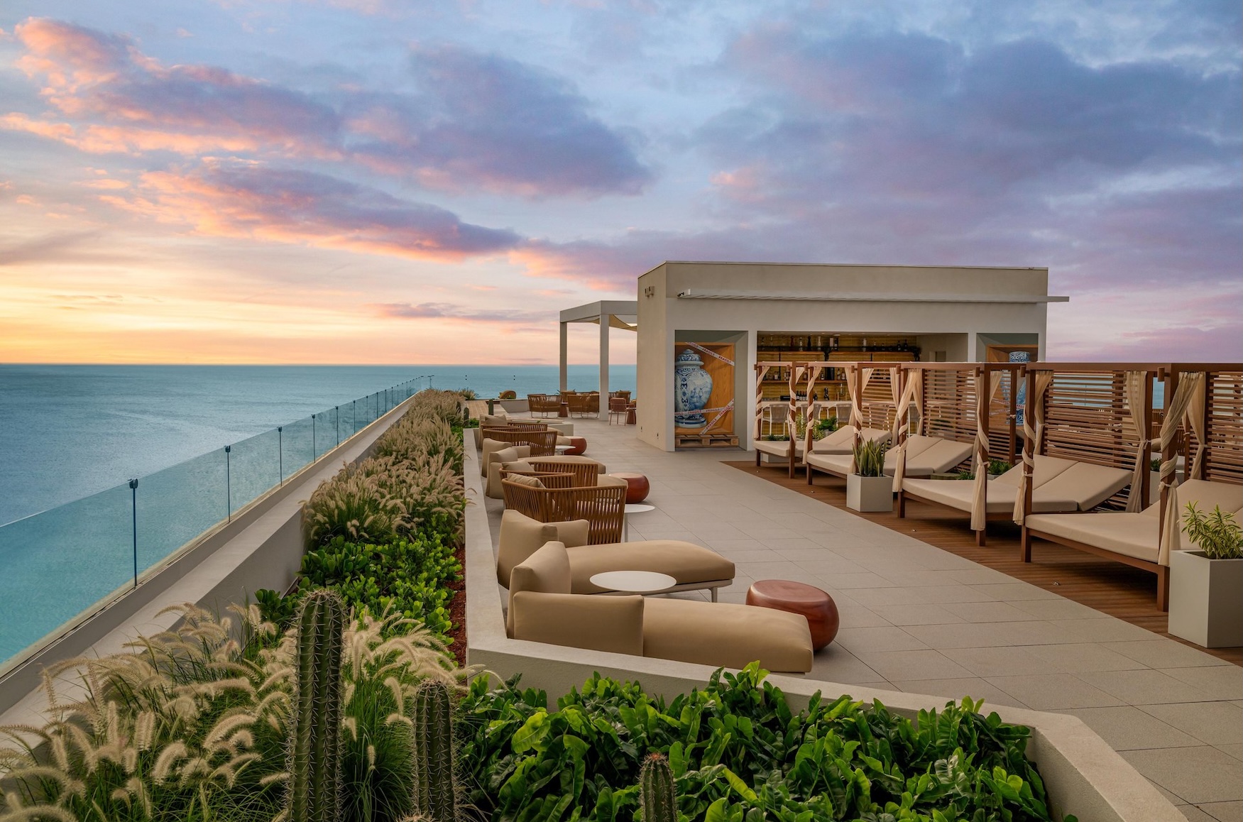 Rooftop lounge with modern seating, cabanas, and greenery overlooks the ocean at sunset under a colorful sky. Glass railing lines the edge, providing unobstructed sea views.