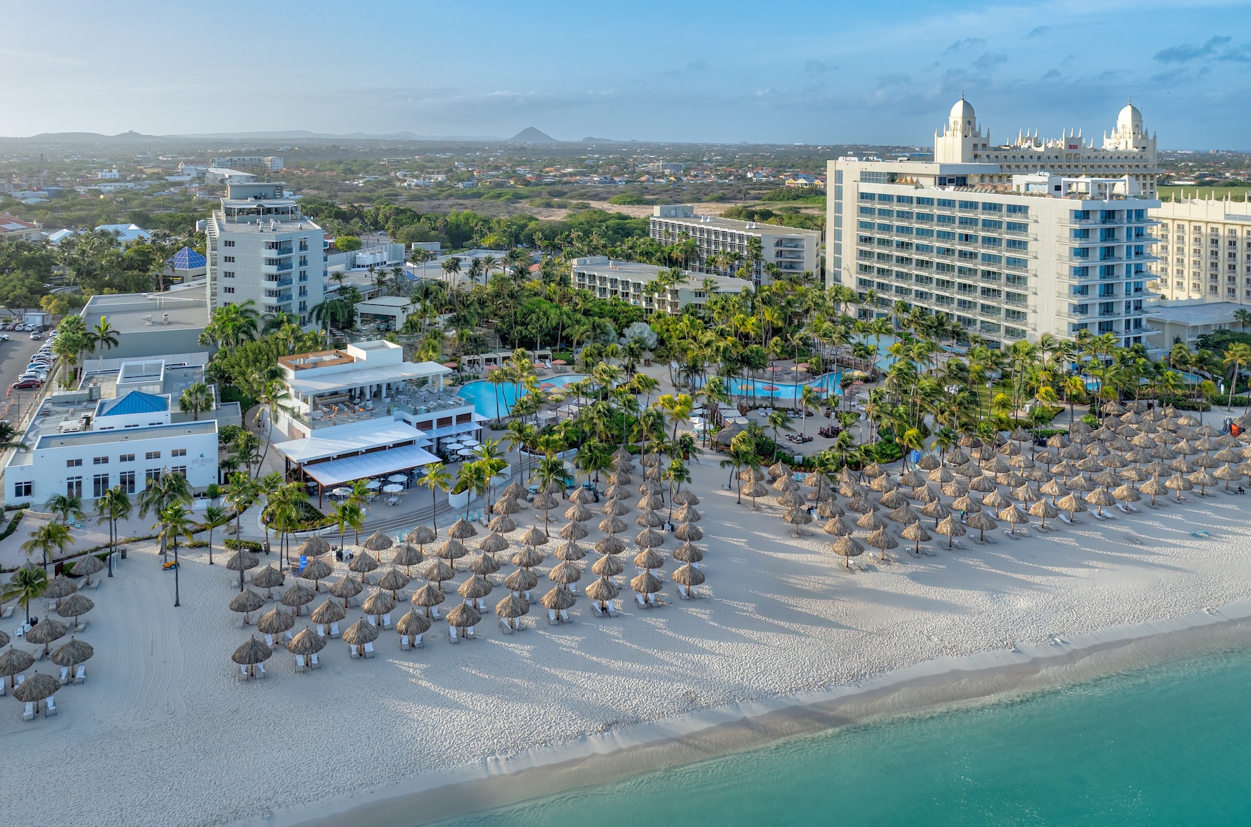 A beachfront resort with tall hotels, pools, palm trees, and rows of thatched umbrellas on white sand, next to clear blue ocean water. The background shows a cityscape and distant hills.