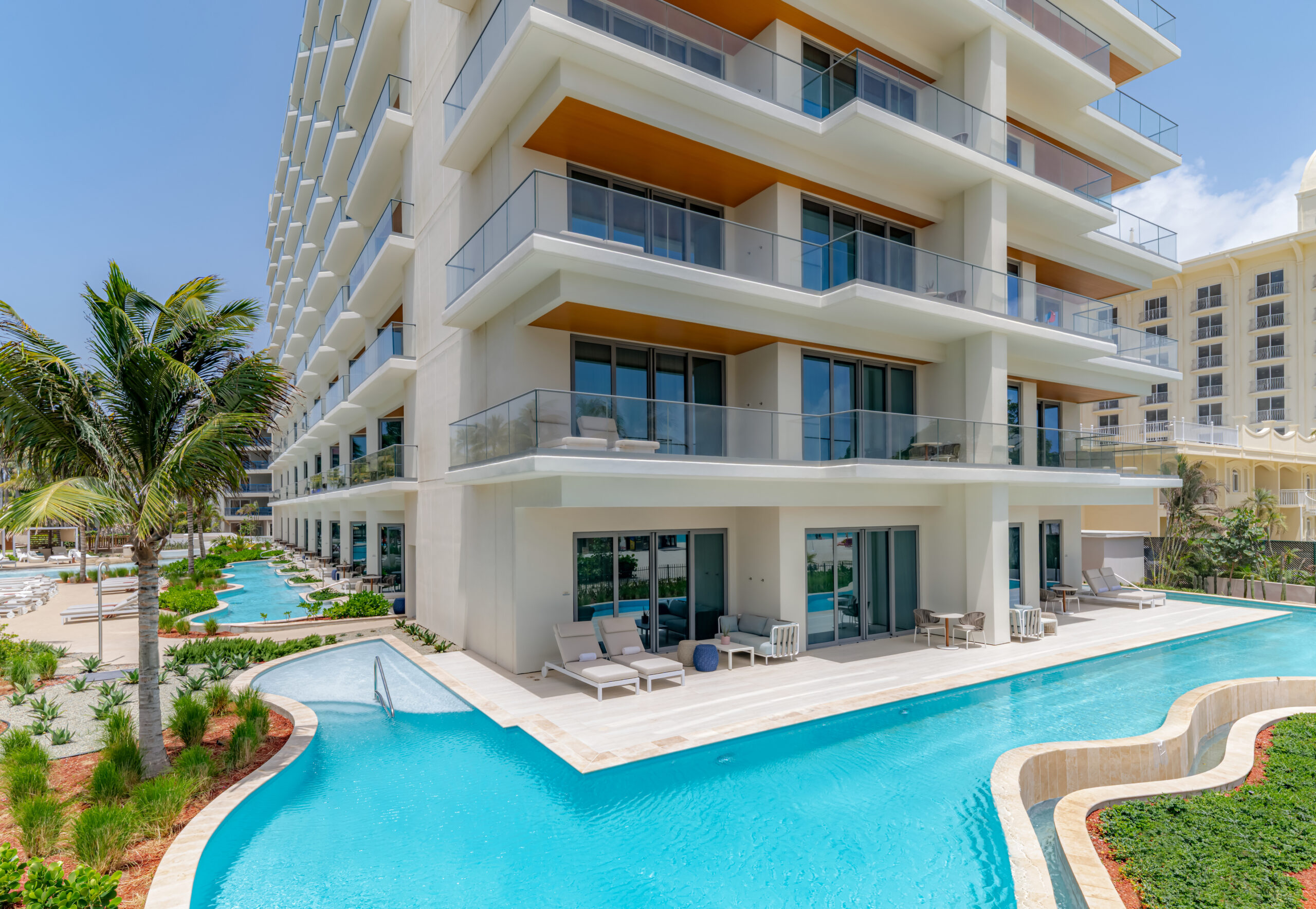 Modern white hotel with private balconies and a winding pool surrounding the ground floor. Palm trees and lounge chairs line the area, under a bright blue sky.
