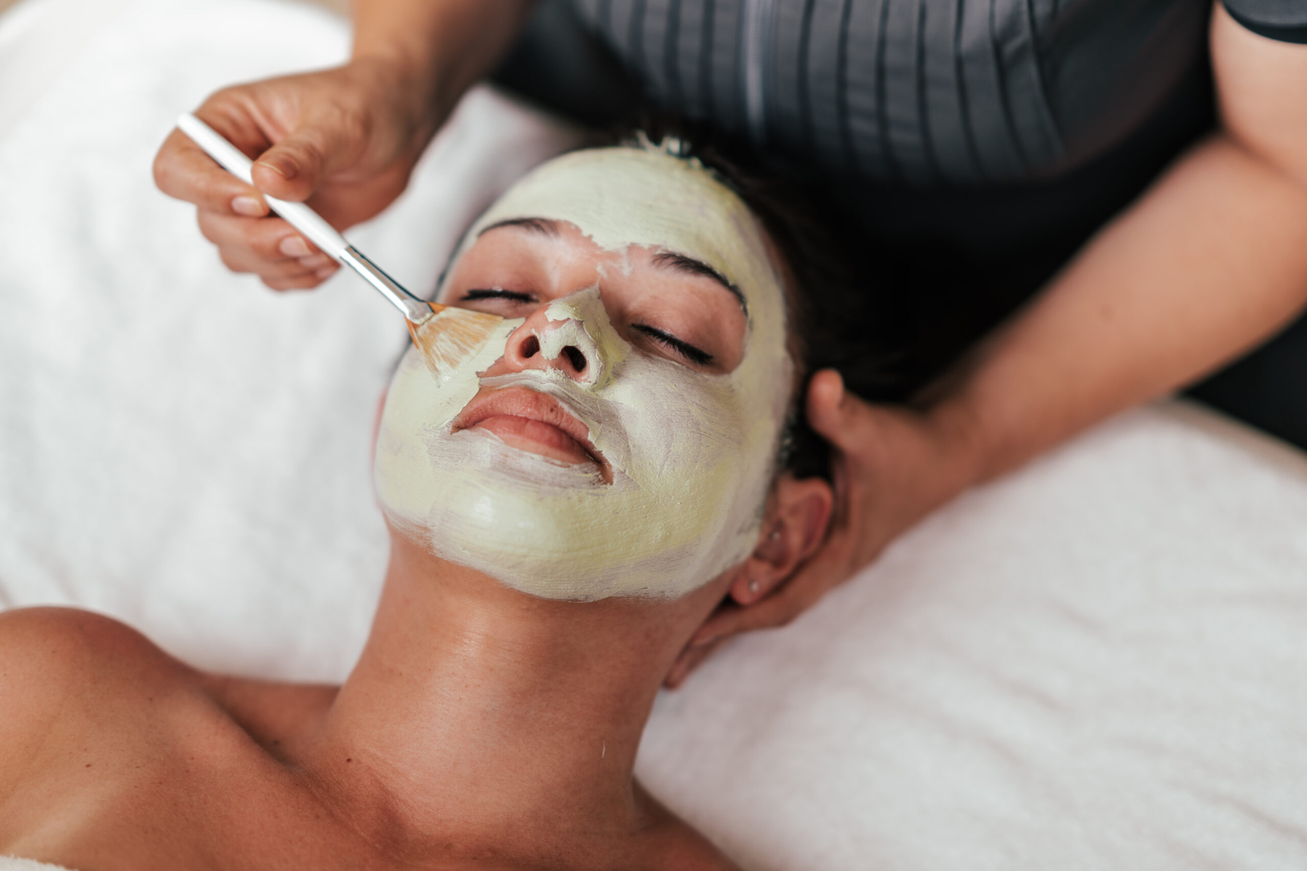 A person receives a facial treatment with a brush applying a green clay mask to their face.
