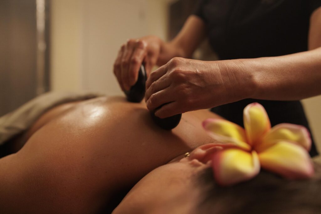 A massage therapist uses smooth, heated stones on a client’s back, with a plumeria flower placed near the client’s ear.