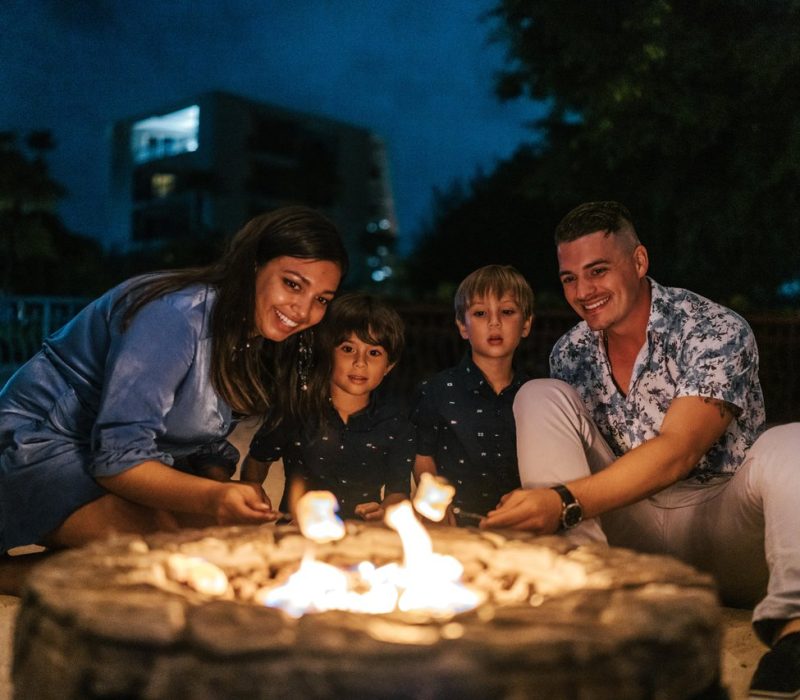 A smiling family of four sits on sand around a campfire at night, roasting marshmallows. There are trees and a building in the background, and everyone appears happy and relaxed.