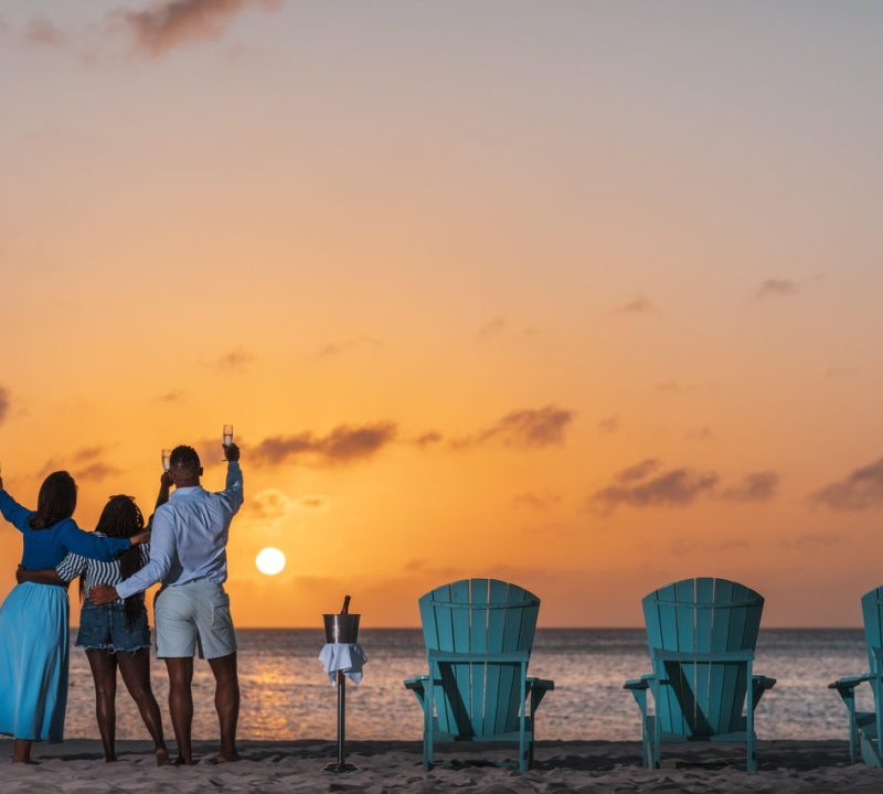 Three people stand on a beach at sunset, raising glasses while facing the ocean. Four blue chairs and a wine cooler sit on the sand, creating a relaxed, celebratory atmosphere.