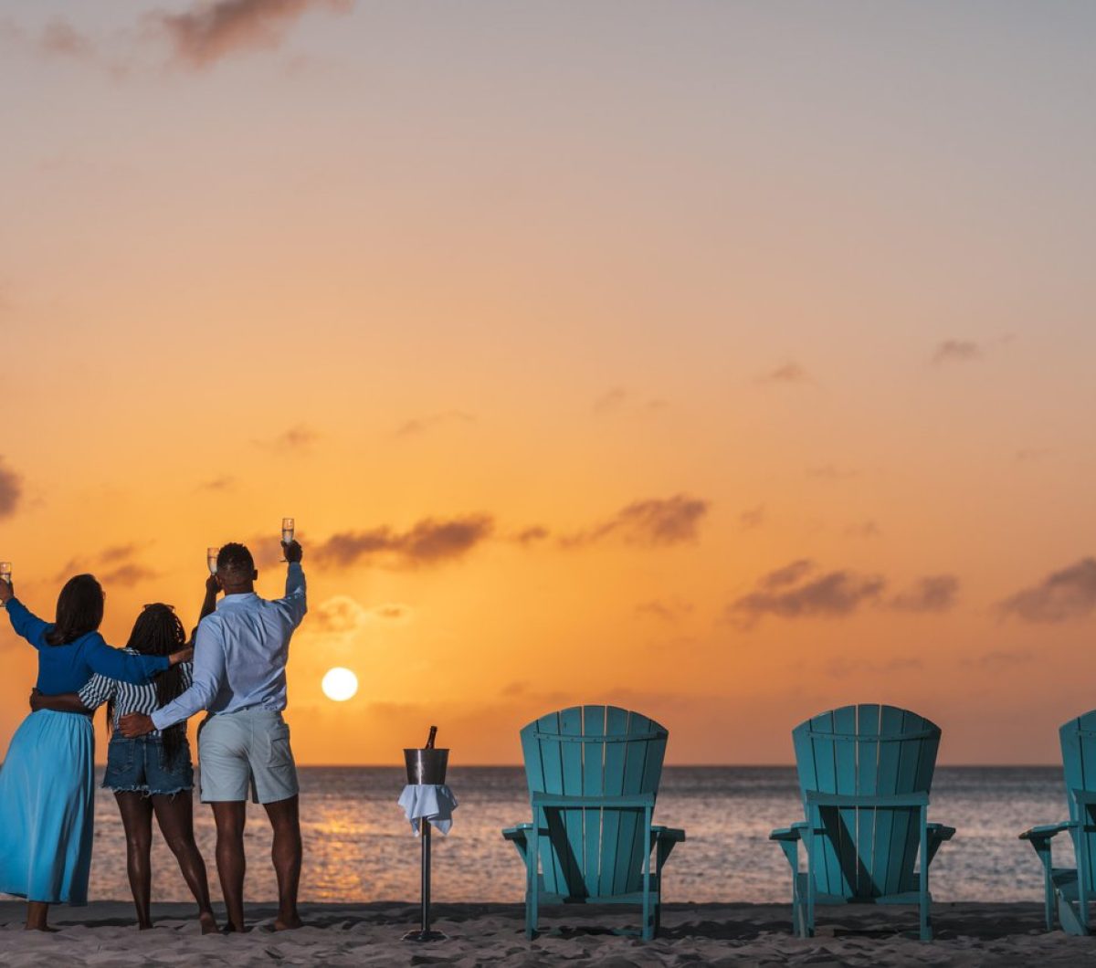 Three people stand on a beach at sunset, raising glasses while facing the ocean. Four blue chairs and a wine cooler sit on the sand, creating a relaxed, celebratory atmosphere.