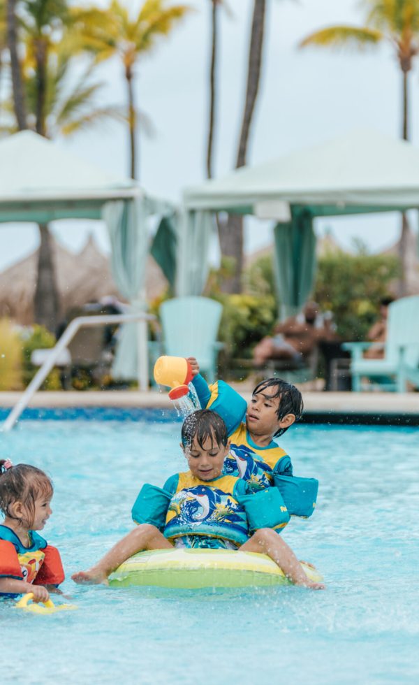 Three young children wearing floaties play together in a swimming pool, splashing water and laughing. Poolside cabanas, palm trees, and lounge chairs are visible in the background on a sunny day.