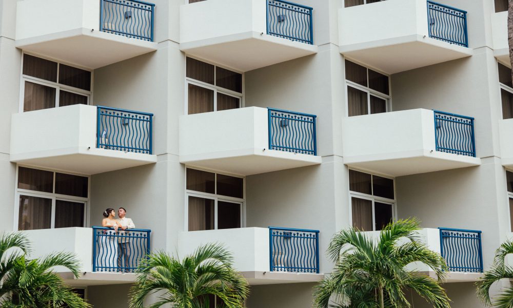 Two people stand together on a balcony of a modern building with multiple white balconies and blue railings; palm trees are visible in front of the building.