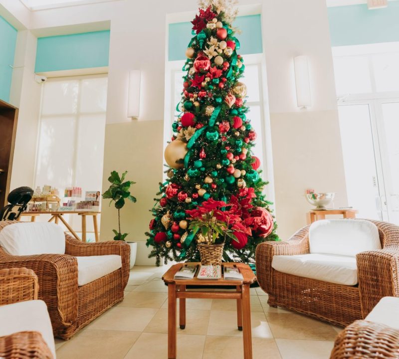 A tall, decorated Christmas tree stands in the center of a bright, modern room with wicker armchairs, white cushions, a small wooden table, potted plants, and festive decorations. Sunlight streams through large windows.