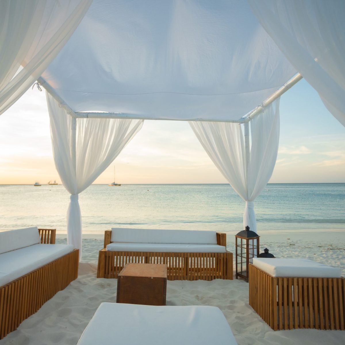 A white canopy with sheer curtains shelters wooden lounge furniture on a sandy beach, overlooking a calm sea at sunset, with sailboats visible in the distance.