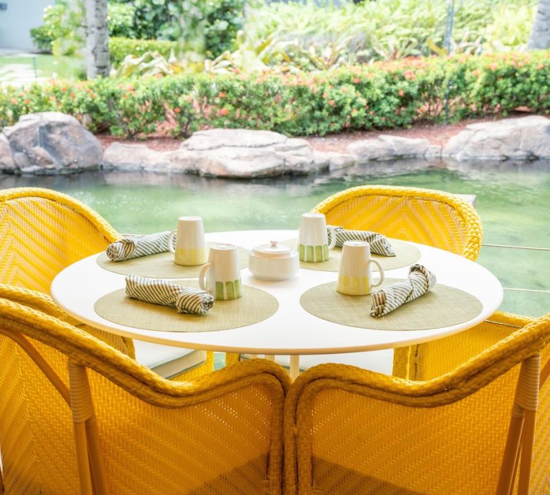 A round white table with four yellow wicker chairs is set with placemats, mugs, napkins, and a teapot, overlooking a lush garden and a pond with rocks.