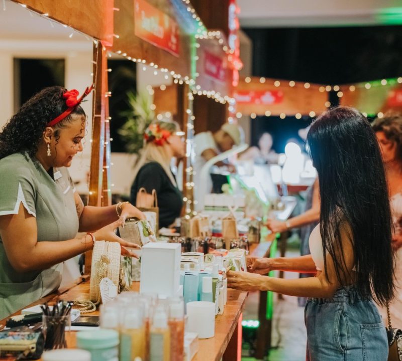 A woman with curly hair and a red headband smiles as she helps a customer at a festive market stall filled with various goods. Other shoppers browse nearby under string lights in a lively indoor setting.