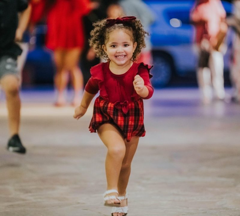 A young girl in a red dress with a plaid skirt and a matching bow runs toward the camera, smiling joyfully. Other people and blurred lights are visible in the background.