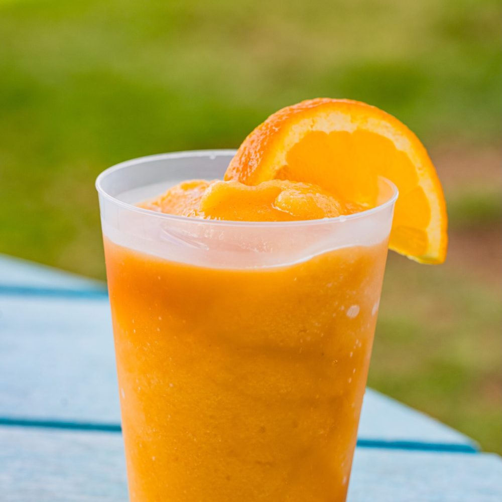 A plastic cup filled with bright orange frozen slush, garnished with a fresh orange slice, sits on a blue wooden surface outdoors with green grass in the background.