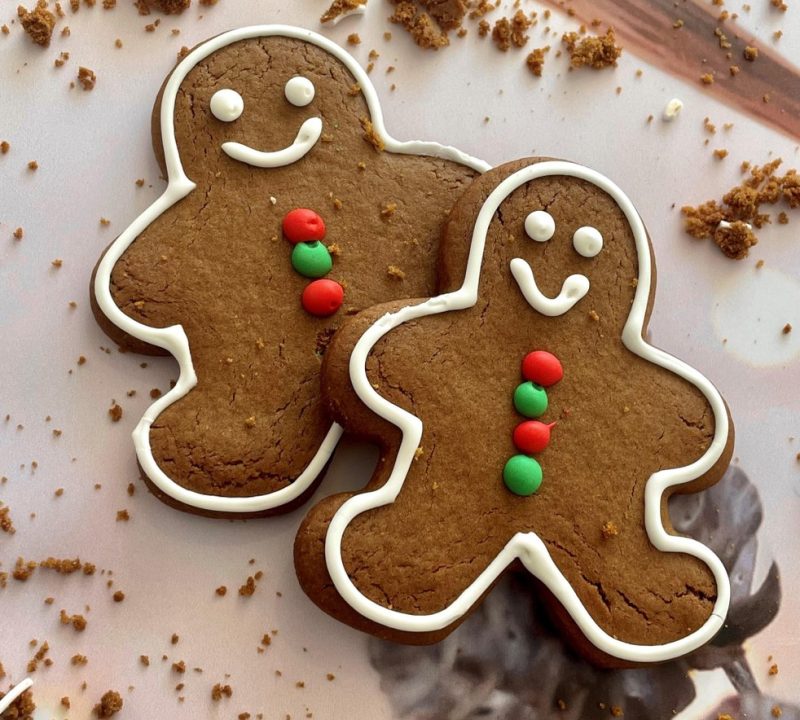 Two decorated gingerbread cookies shaped like people, each with white icing smiles and buttons made of red and green candies, lie on a surface scattered with cookie crumbs.