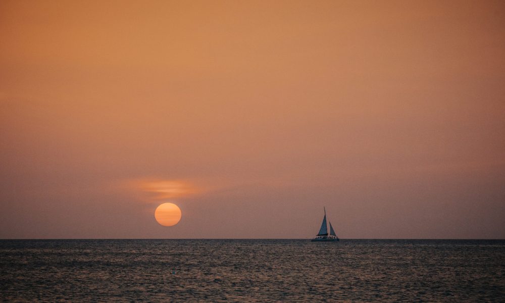 A sailboat floats on calm ocean waters during sunset, with the sun low on the horizon and the sky glowing in warm orange tones.