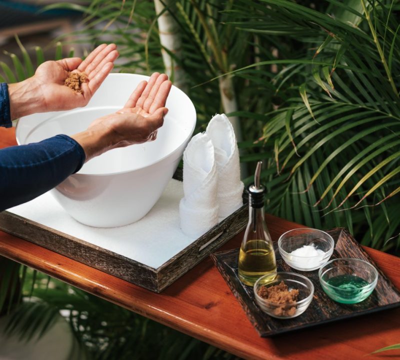 A person holding brown scrub in their hands stands near a white bowl and tray with small bowls of oil, sugar, brown scrub, and green gel, surrounded by lush green plants.