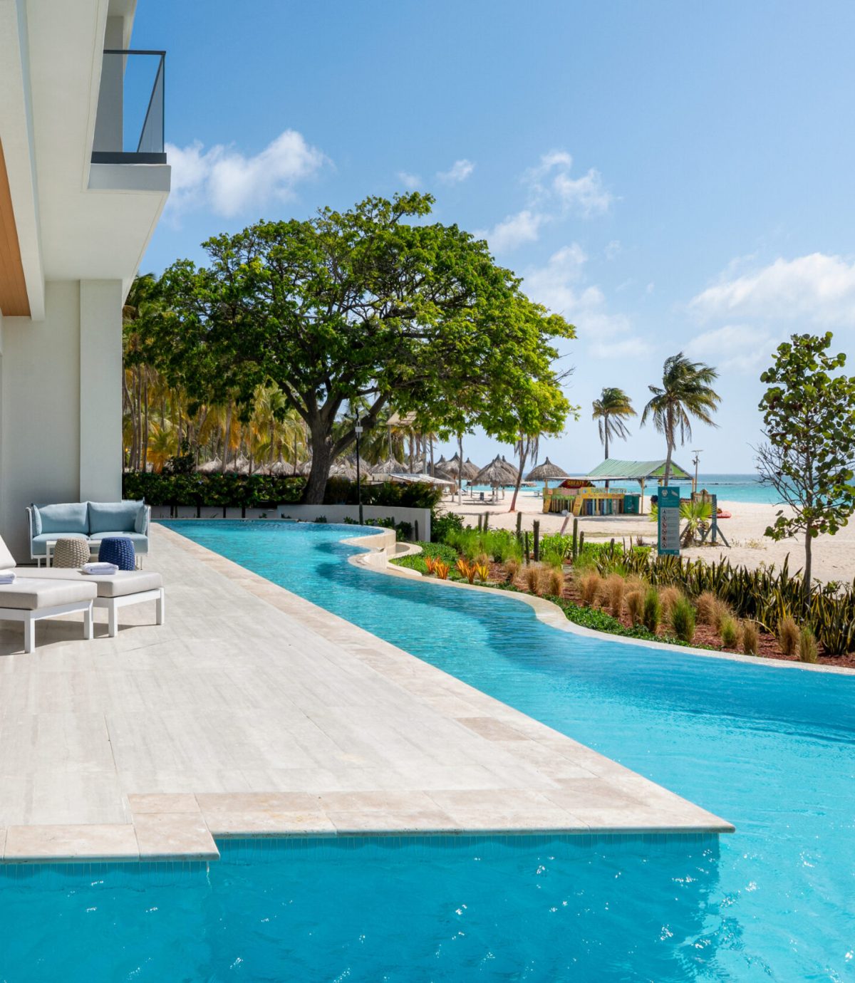 Modern beachfront patio with lounge chairs beside a private pool, overlooking sandy beach, palm trees, and clear blue sky. The ocean is visible in the background, creating a relaxing tropical atmosphere.