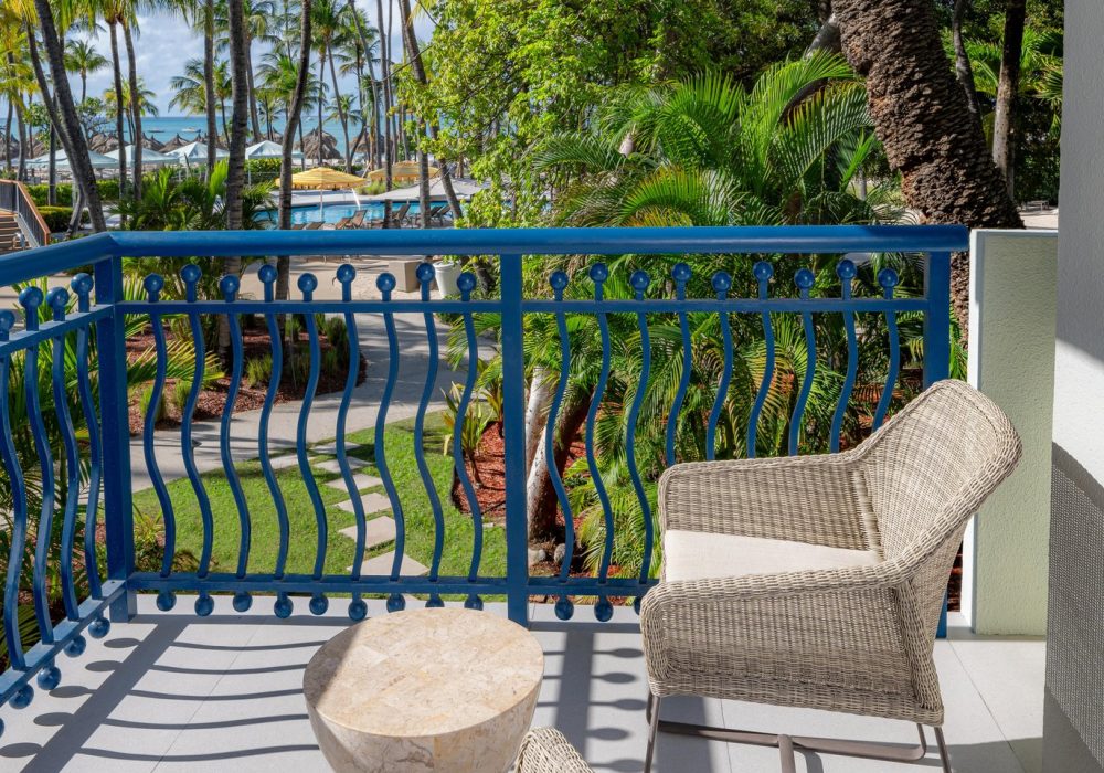 A balcony with two wicker chairs and a small stone table overlooks a lush, tropical garden with palm trees and a path leading toward the beach under a sunny sky.