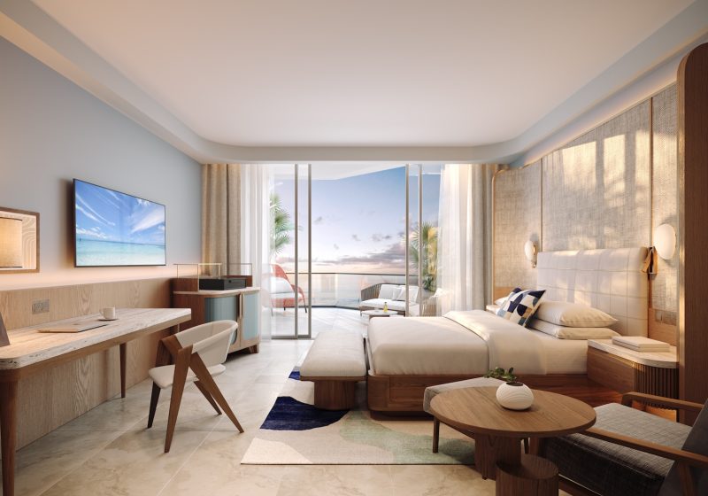 A modern hotel room with a large bed, desk, TV, and cozy seating area. Glass doors open to a balcony overlooking the ocean at sunset, with warm lighting and stylish, contemporary decor throughout the space.