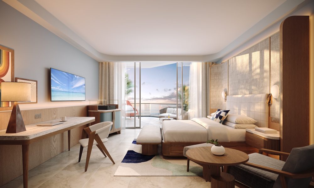 A modern hotel room with a large bed, desk, TV, and cozy seating area. Glass doors open to a balcony overlooking the ocean at sunset, with warm lighting and stylish, contemporary decor throughout the space.