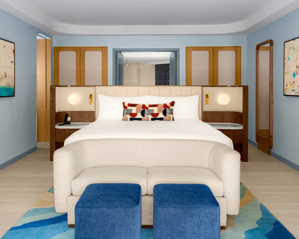 Modern hotel room with a king bed, geometric pillows, light sofa, two blue ottomans, abstract art on blue walls, and wooden accents, creating a clean, stylish, and inviting atmosphere.
