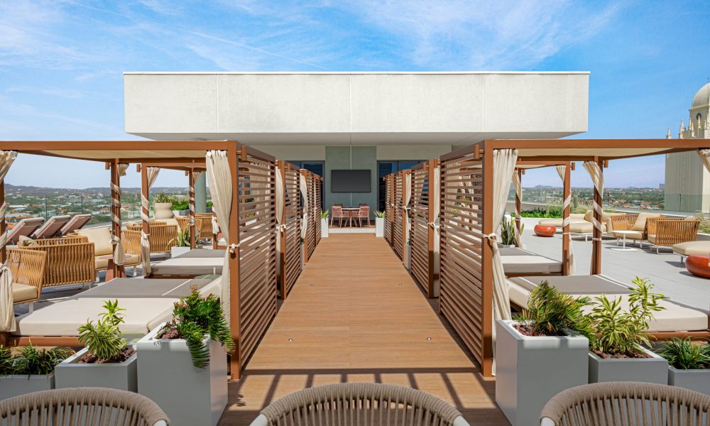 Rooftop lounge with wooden cabanas, cushioned seating, potted plants, and clear blue sky. Outdoor furniture and a walkway lead to a shaded area at the far end.