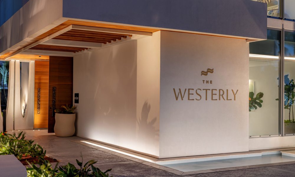 Modern building entrance at dusk with “The Westerly” sign on a beige wall, wooden ceiling accents, potted plant, and glass windows. Subtle lighting highlights the clean, upscale design and surrounding greenery.