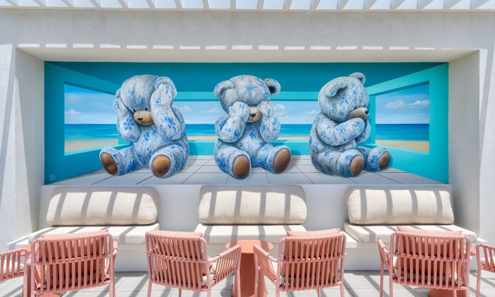 A mural of three blue teddy bears in “see no evil, hear no evil, speak no evil” poses sits behind cream sofas and pink chairs, with an ocean view painted in the background.