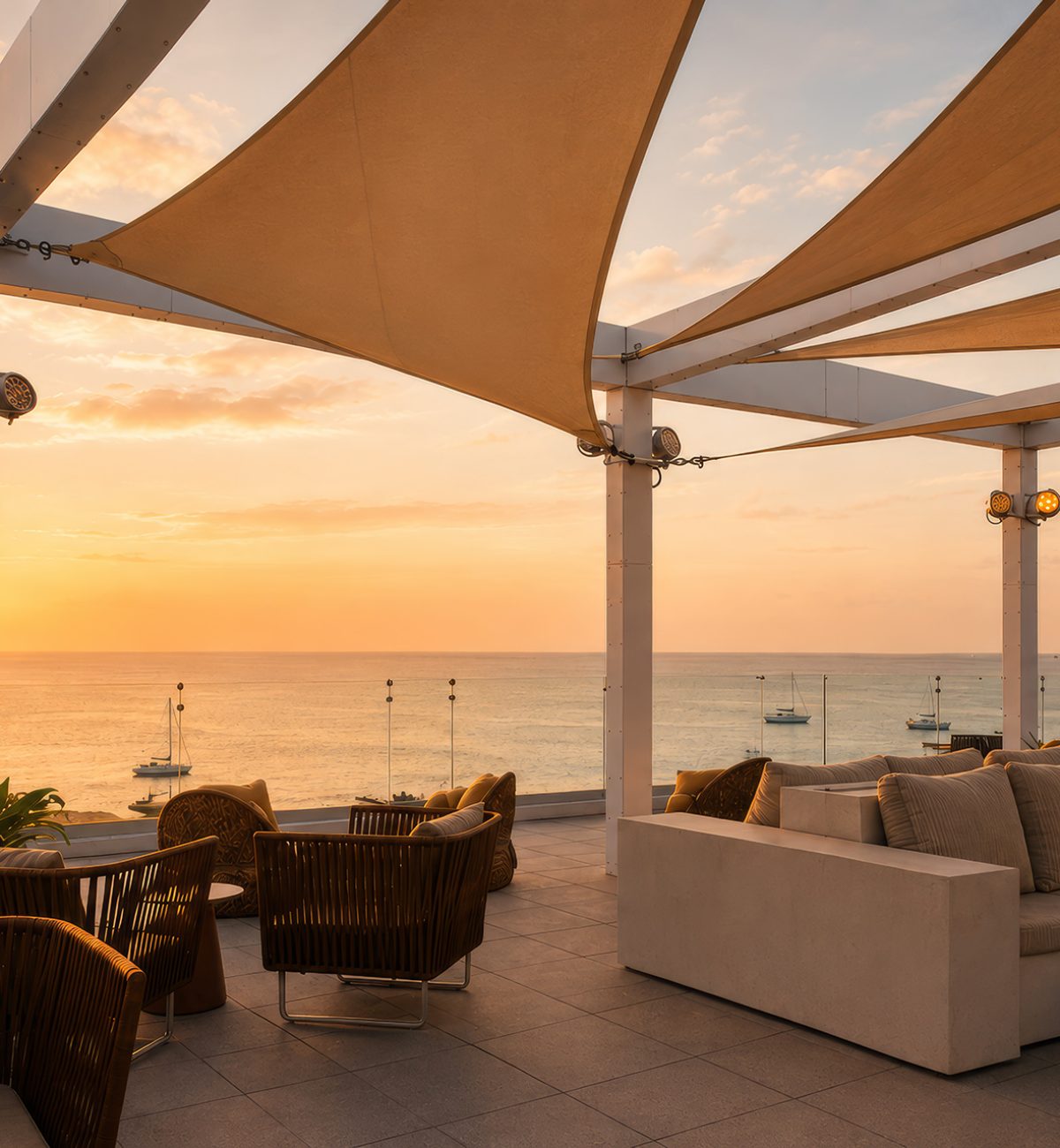 Rooftop lounge with modern sofas and chairs under fabric canopies, overlooking the ocean at sunset. Several boats are visible on the calm water, with a colorful sky in the background.